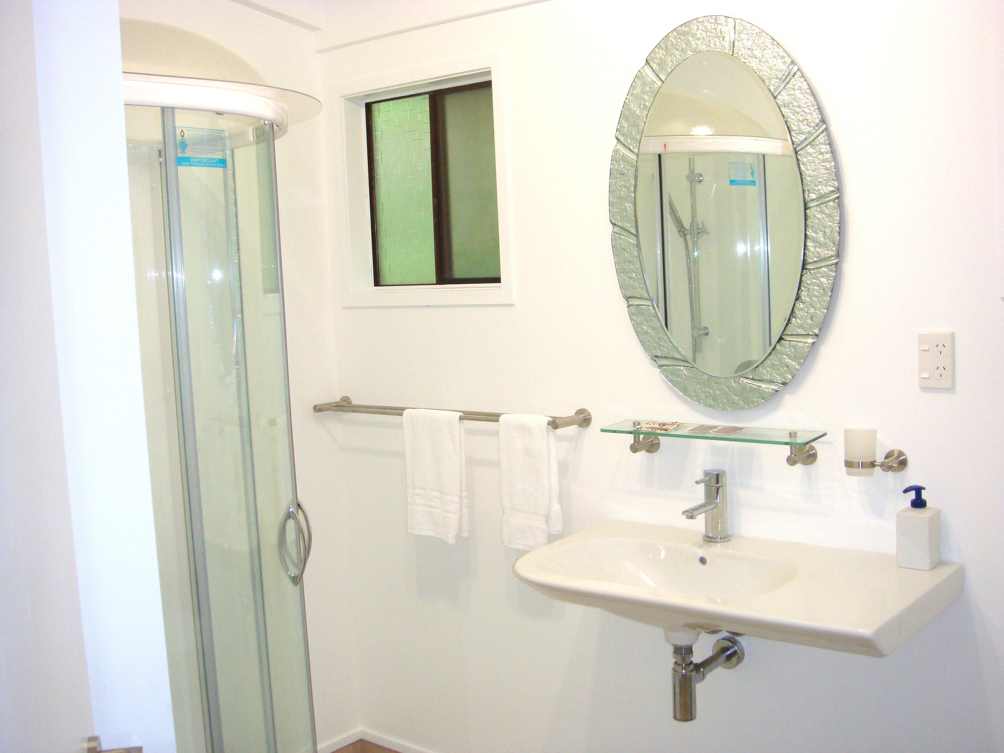 A bathroom with a glass shower enclosure, a white sink with a glass shelf, and a mirror with an ornate frame.