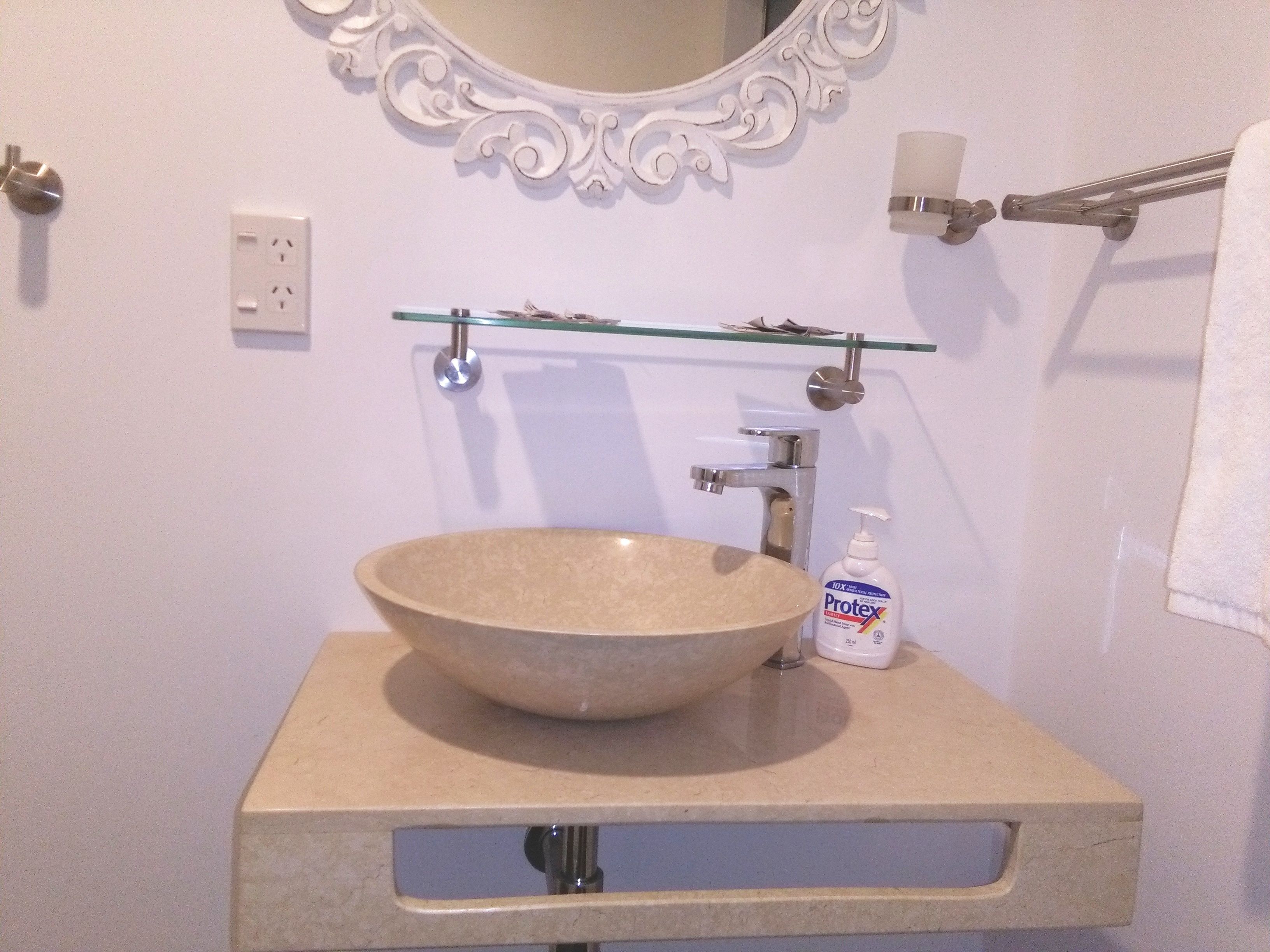 Een badkamer met een beige wastafel, een glazen plank erboven, een witte zeepdispenser en een spiegel met decoratieve details.