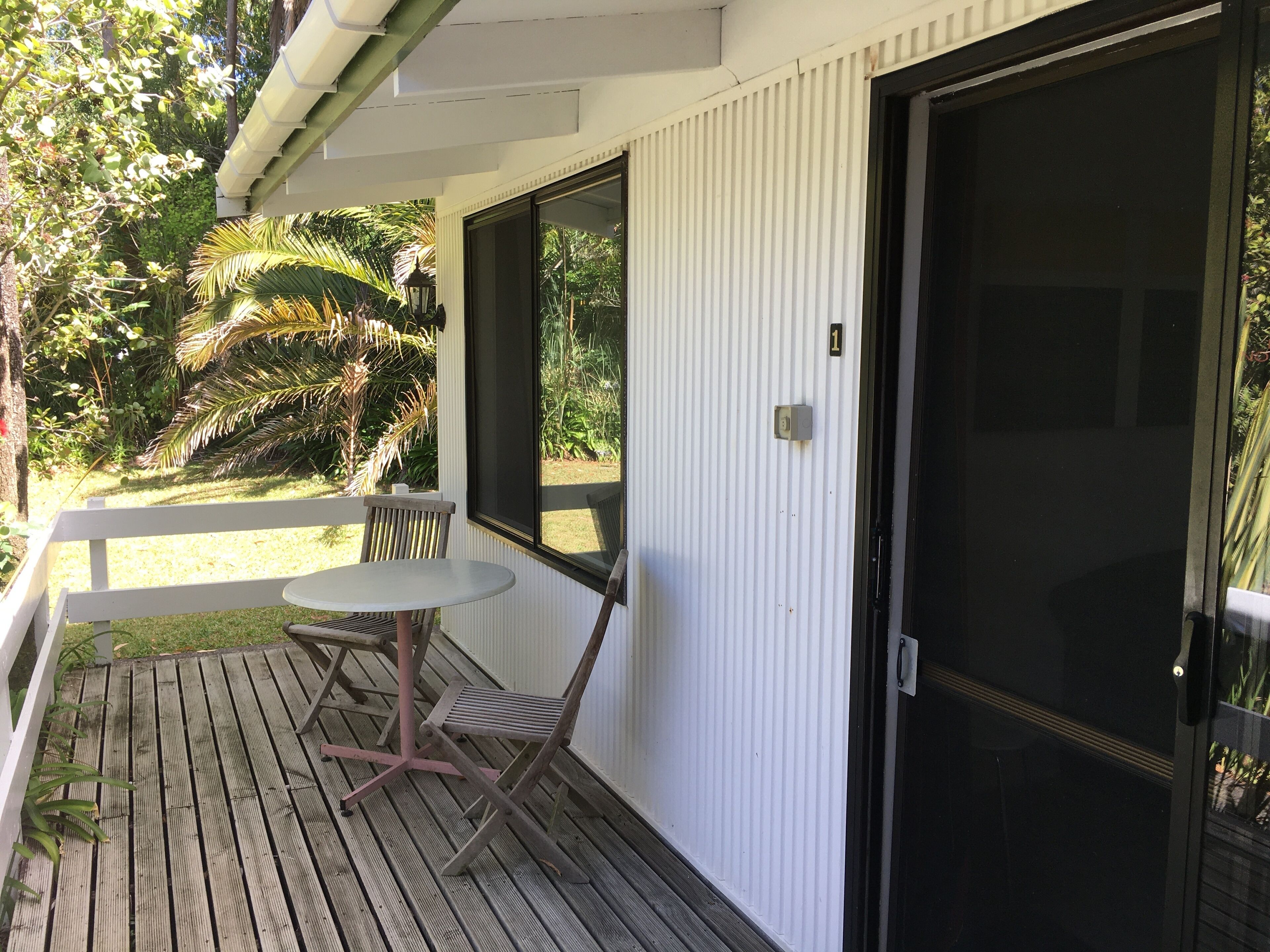 A wooden deck with a table and chairs, a door, and a view of a garden with palm trees.