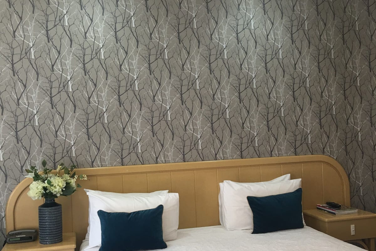 A hotel room with a bed, two pillows, a nightstand with a vase of flowers, and a wall with a tree pattern.