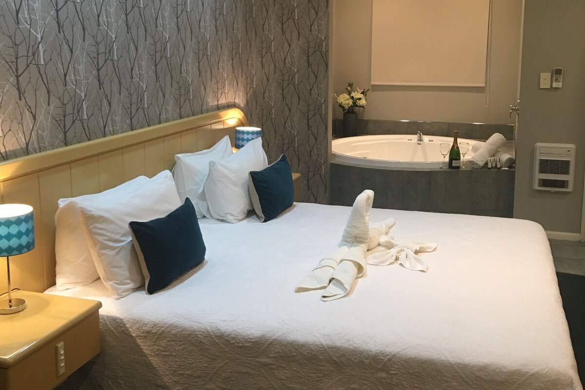 A hotel room with a large bed, a nightstand, a lamp, a wall-mounted air conditioner, a mirror, a bathroom with a bathtub, and a towel rack.