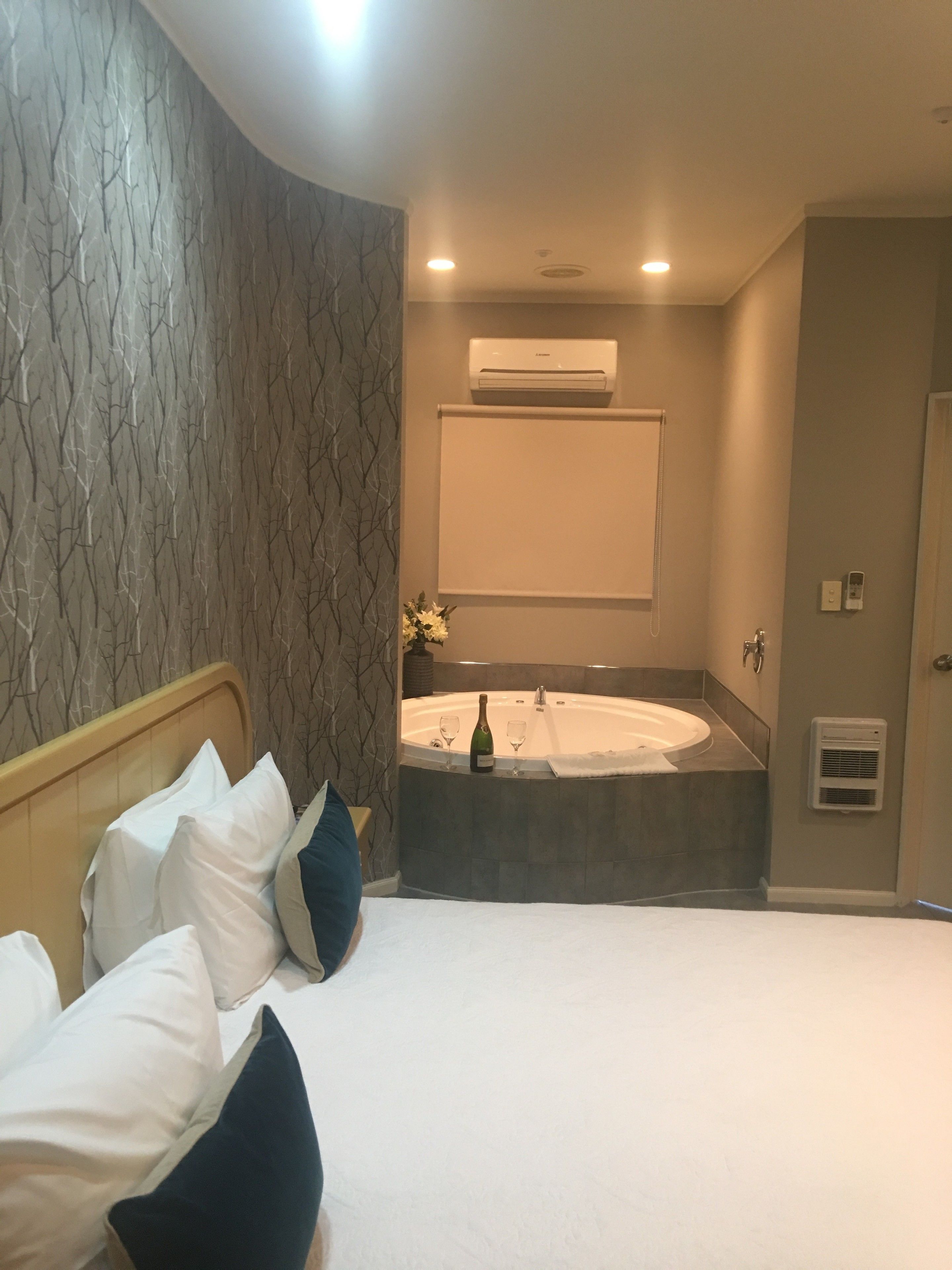 A hotel room with a bed, a large window, a wall-mounted air conditioner, and a bathroom with a bathtub and a towel rack.