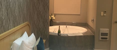 A hotel room with a bed, a large window, a wall-mounted air conditioner, and a bathroom with a bathtub and a towel rack.