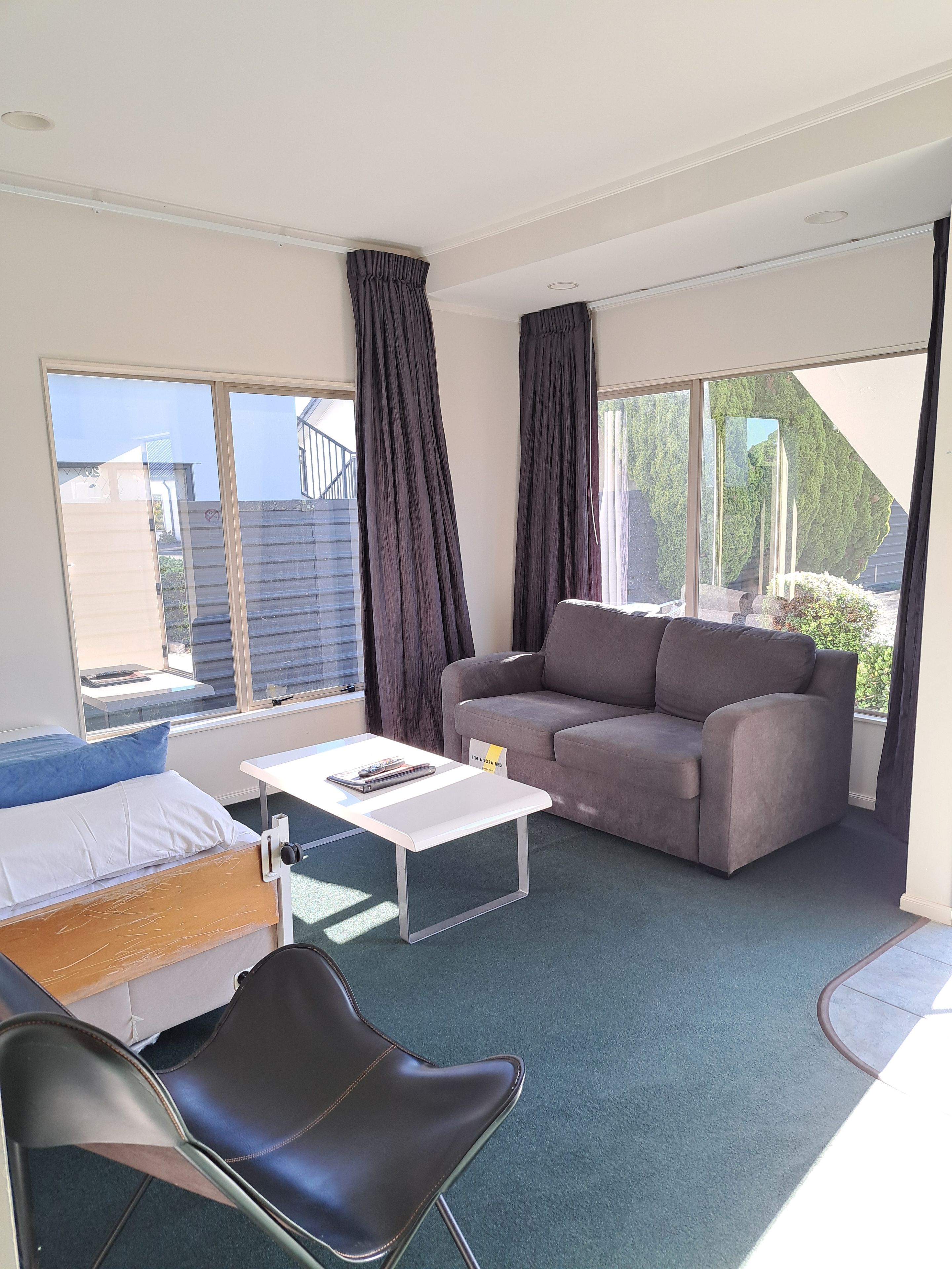 Una habitación con una cama, un sofá, una mesa de centro y una silla. Amplias ventanas con cortinas.