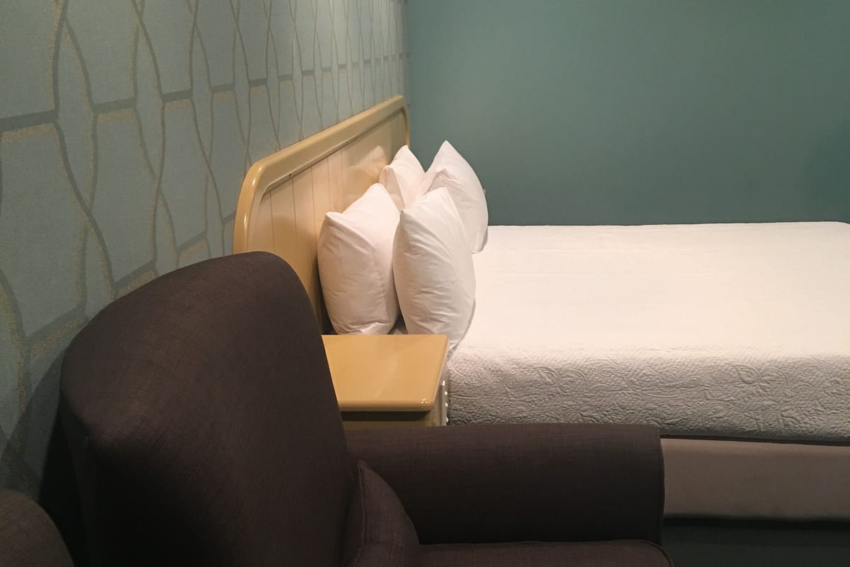 A hotel room with a bed, a wooden nightstand, a brown armchair, and a patterned wall.
