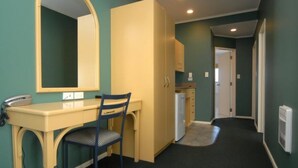 A compact room with a desk, chair, and a large mirror. There is a kitchenette area with cabinets and a refrigerator.
