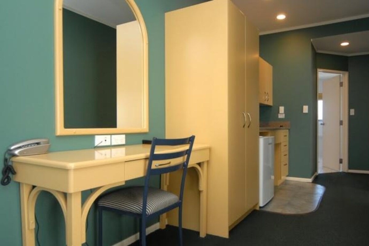 A compact room with a desk, chair, and a large mirror. There is a kitchenette area with cabinets and a refrigerator.