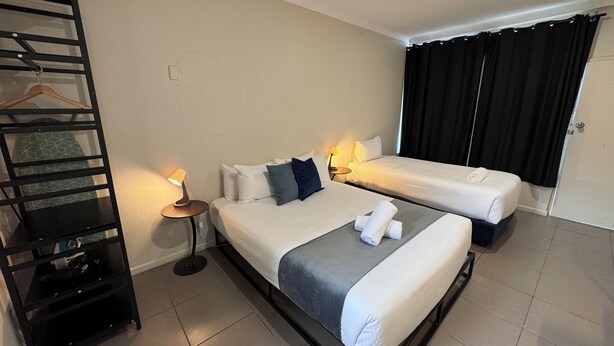 A hotel room with two beds, a black metal shelf unit, a small bedside table with a lamp, and a door with black curtains. - Lismore City Motor Inn (Lismore)