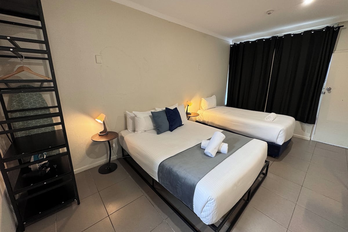 Une chambre d’hôtel avec deux lits, une étagère en métal noir, une petite table de chevet avec une lampe et une porte avec des rideaux noirs.