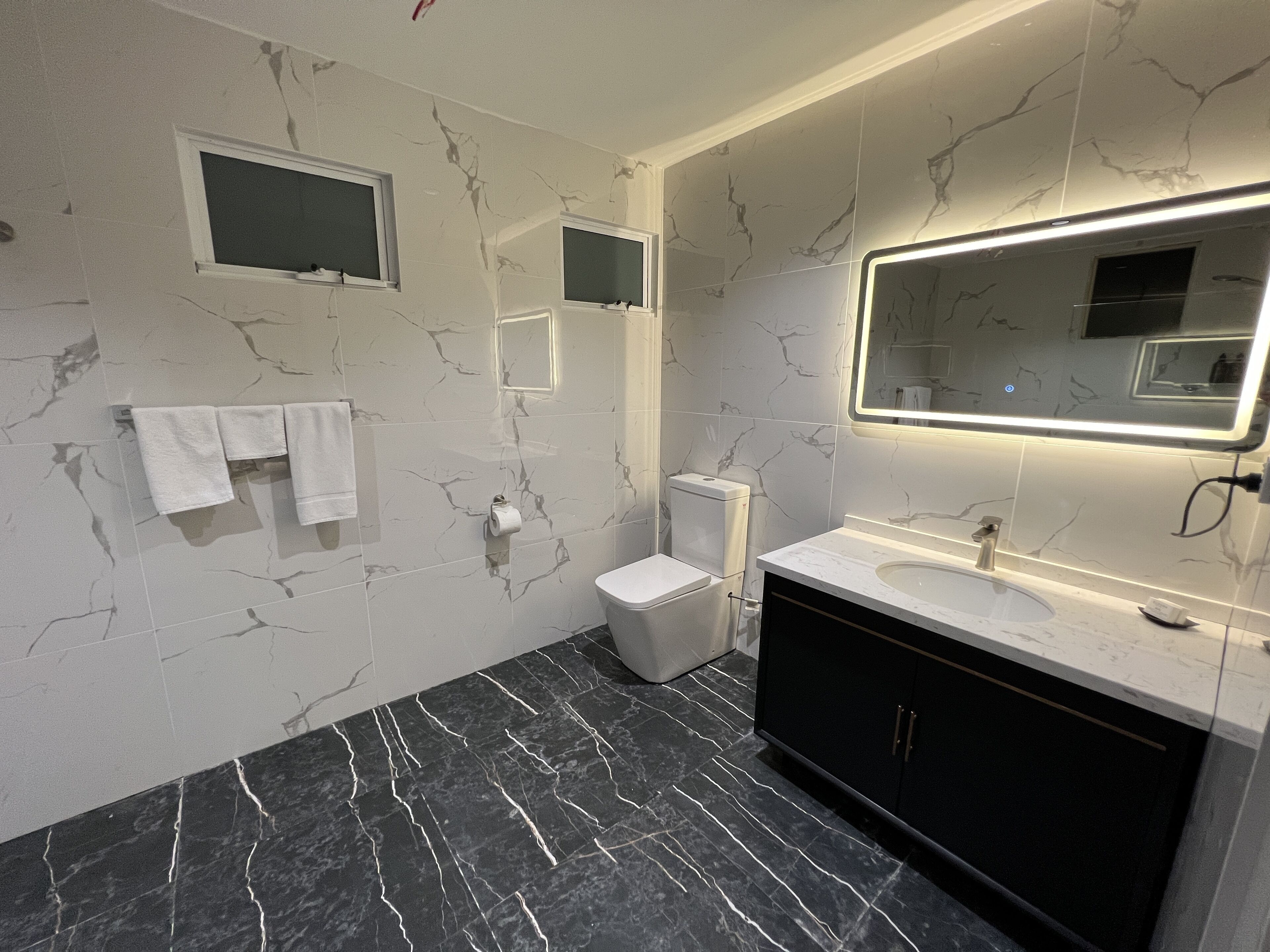 Une salle de bain moderne avec un sol en marbre, des toilettes, un lavabo avec un miroir et deux porte-serviettes.