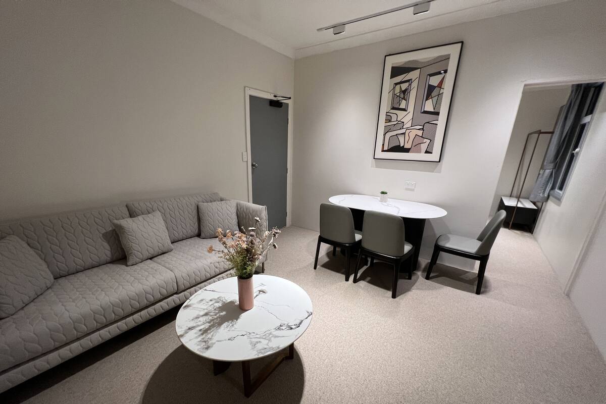 Un salon moderne avec un canapé gris, une table basse ronde ornée d’un vase de fleurs et un coin repas avec une table et des chaises blanches.
