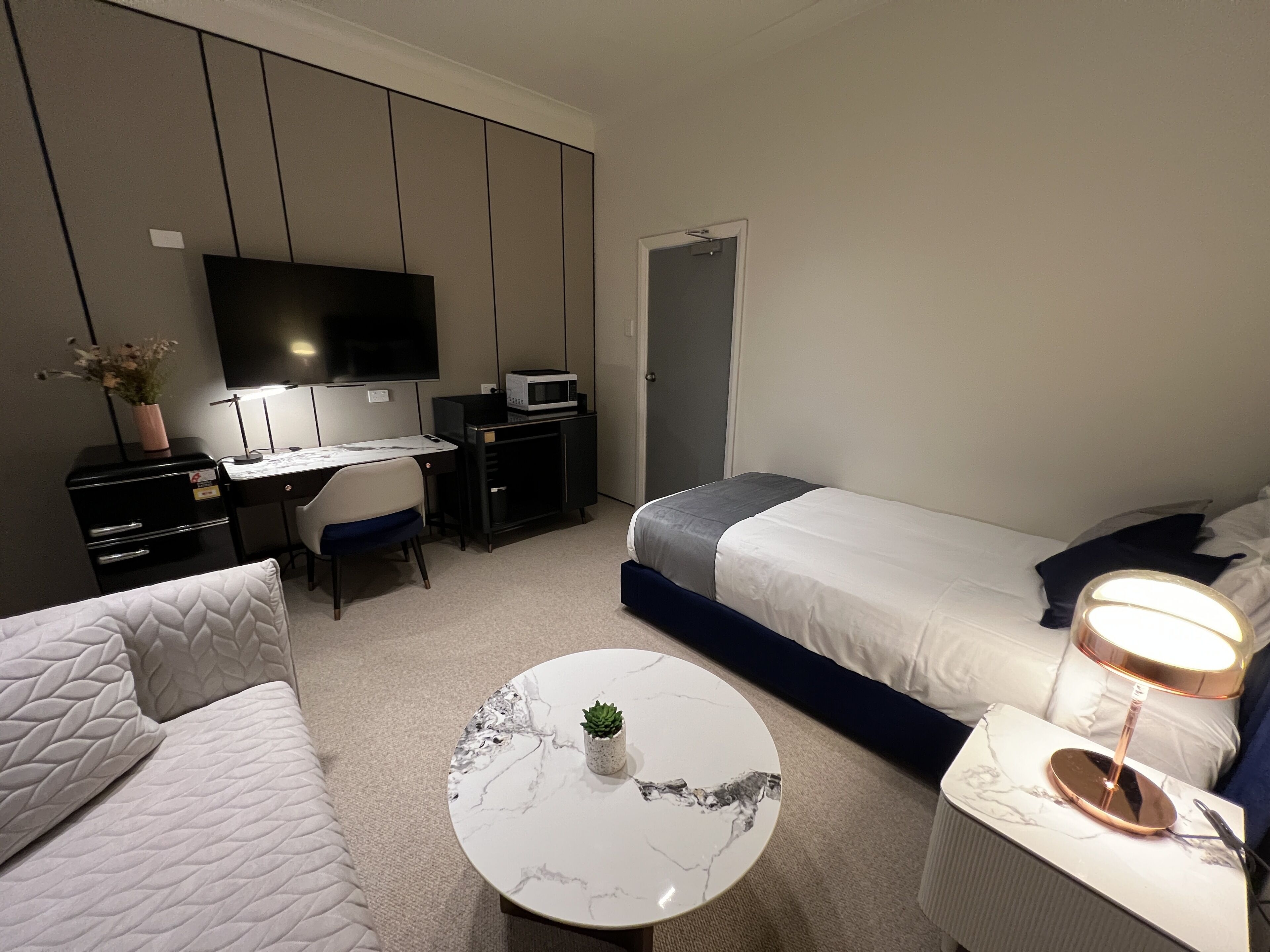 A modern hotel room with a bed, a desk, a chair, a TV, a microwave, a small plant, and a bedside lamp.