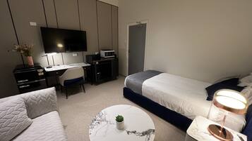 A modern hotel room with a bed, a desk, a chair, a TV, a microwave, a small plant, and a bedside lamp.