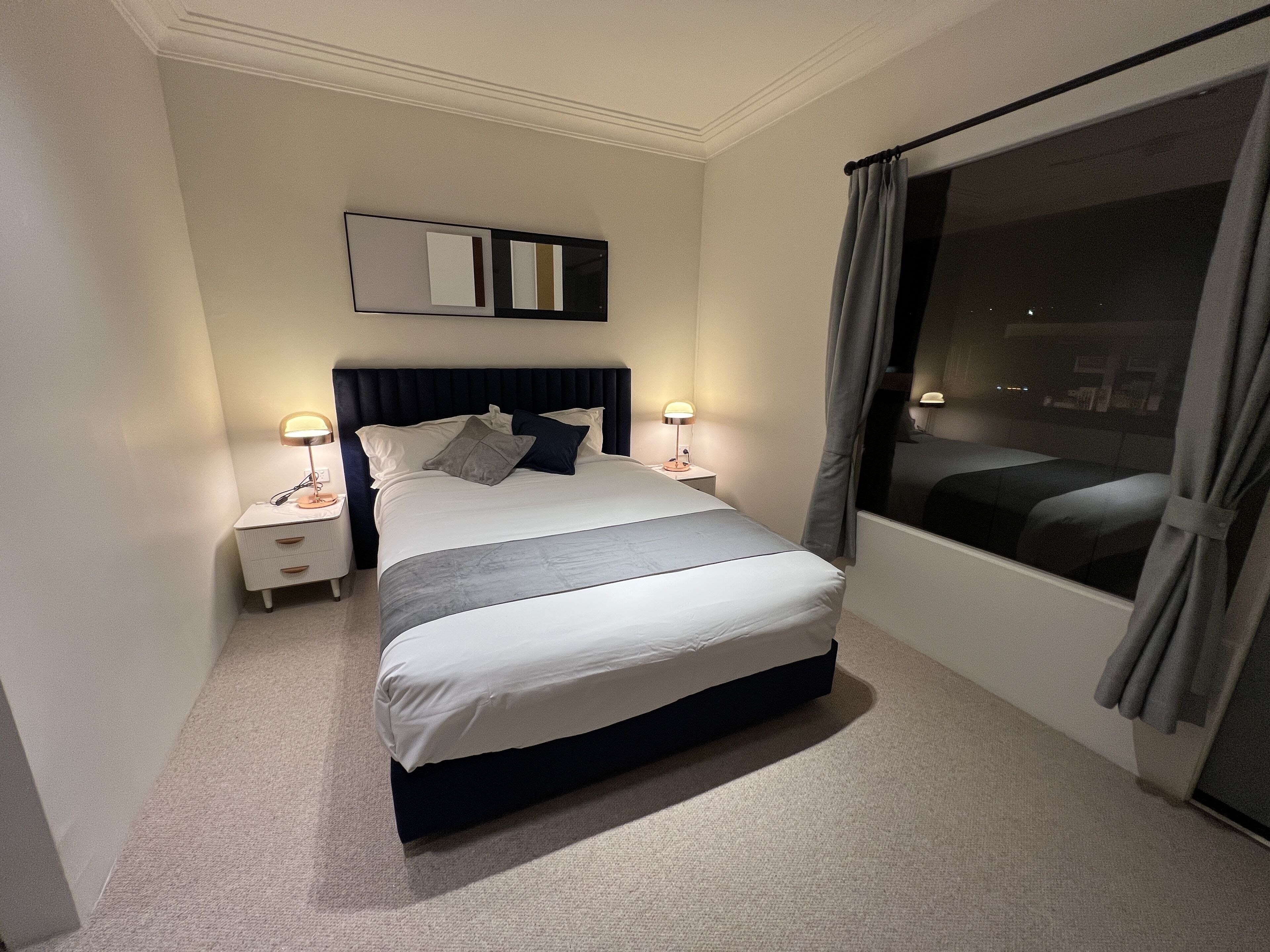 A neatly made bed with a dark headboard, two bedside lamps, and a large window with curtains.