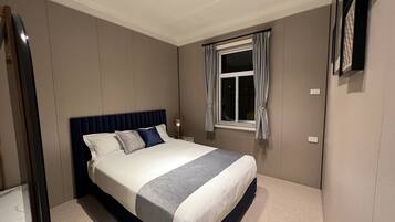 A bedroom with a bed, a nightstand, a window with curtains, and a mirror on the wall.