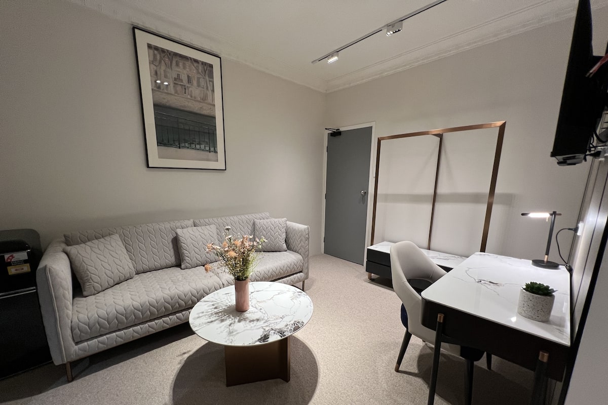 Un salon moderne avec un canapé gris, une table basse ronde, une petite table d’appoint, un bureau avec une chaise et un tableau encadré au mur.