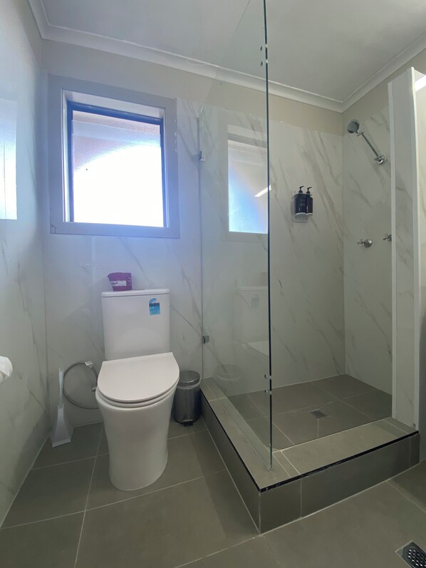 Un baño con inodoro, ducha con cerramiento de vidrio y ventana sobre el inodoro.