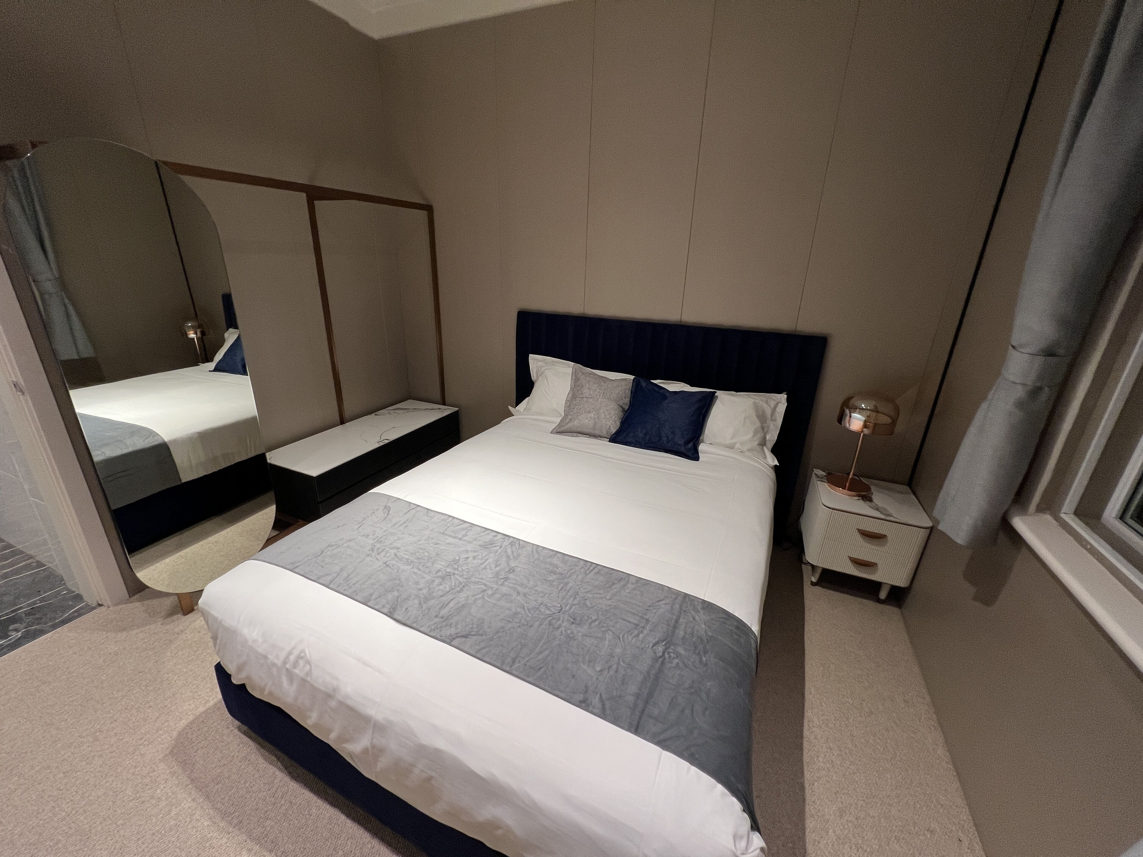 A hotel room with a large bed, a bedside table, a mirror, and a window with curtains.