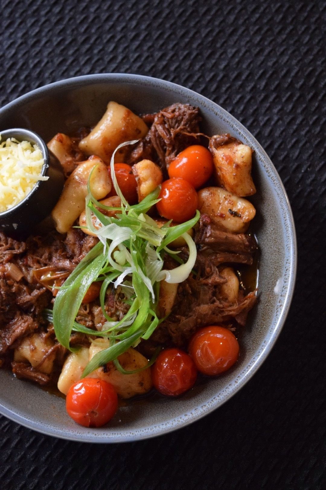A bowl of pulled pork with vegetables and a side of shredded cheese.