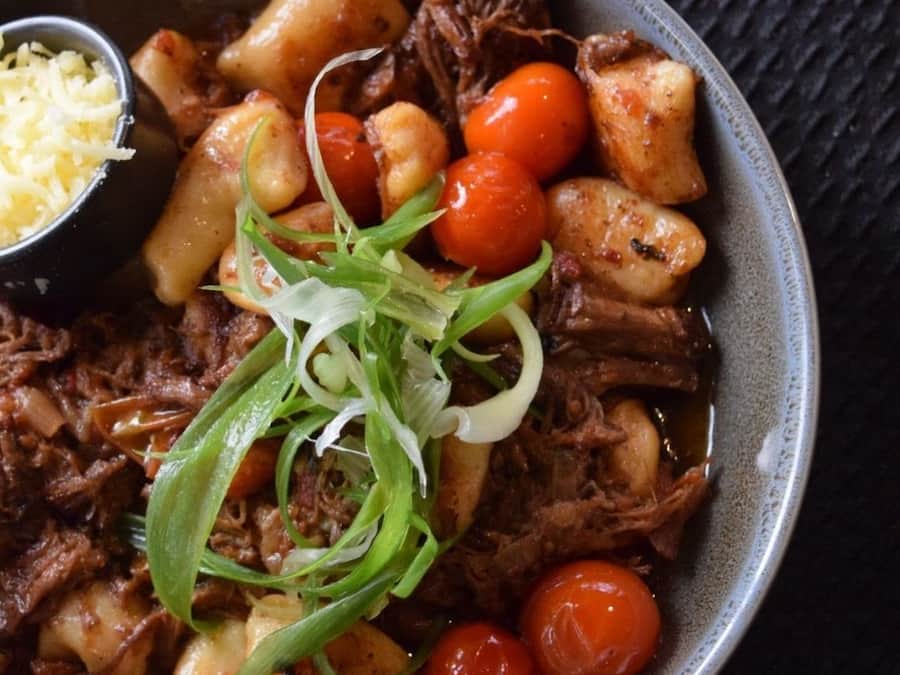 A bowl of pulled pork with vegetables and a side of shredded cheese.