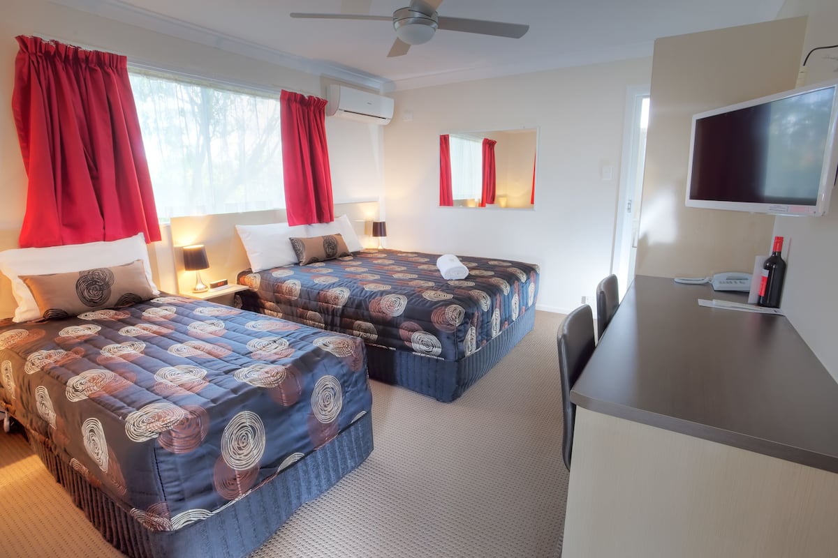 A hotel room with two beds, a desk, a TV, and a window with red curtains.