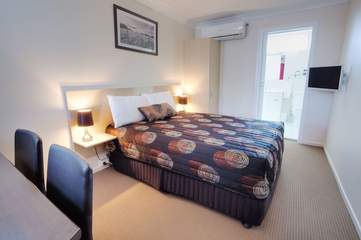 A hotel room with a bed, bedside lamps, a desk with chairs, a framed picture on the wall, and an air conditioning unit.