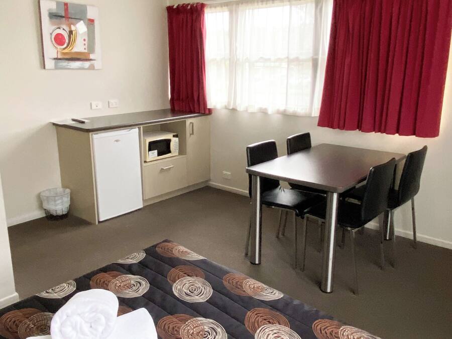 Una habitación compacta con cama, un pequeño rincón americano, una mesa de comedor con sillas y una ventana con cortinas rojas.