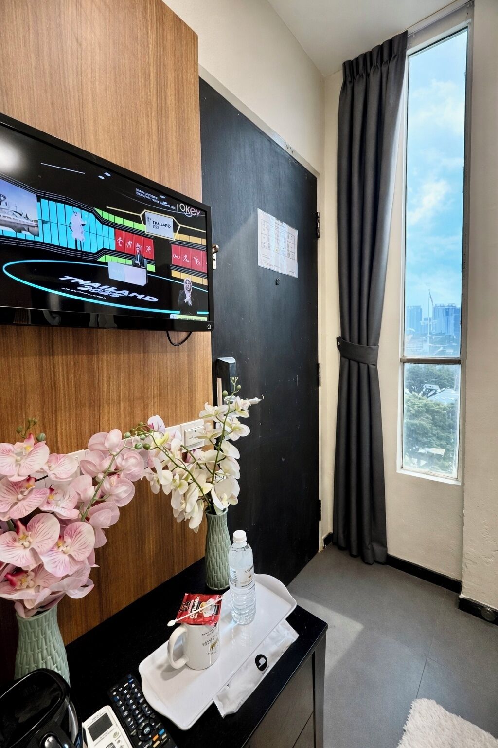 A hotel room with a large window, a flat-screen TV mounted on the wall, a desk with a vase of flowers, a water bottle, a cup, and a remote control.
