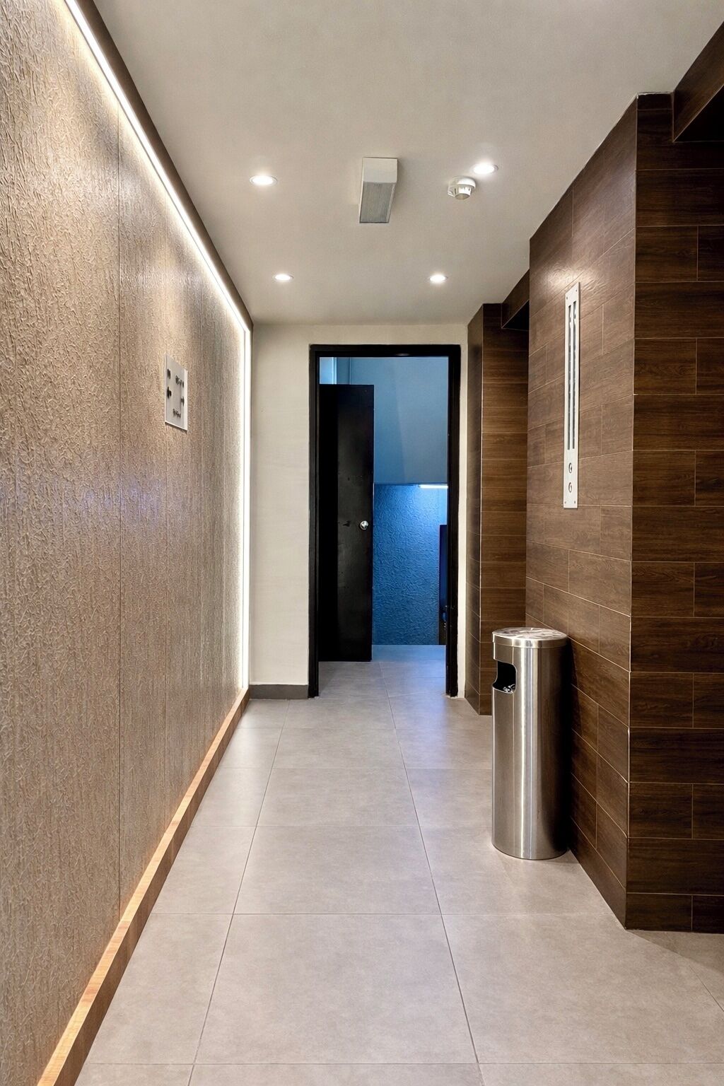 A modern hallway with a light-colored floor, wooden paneling on one side, and a trash can on the other.