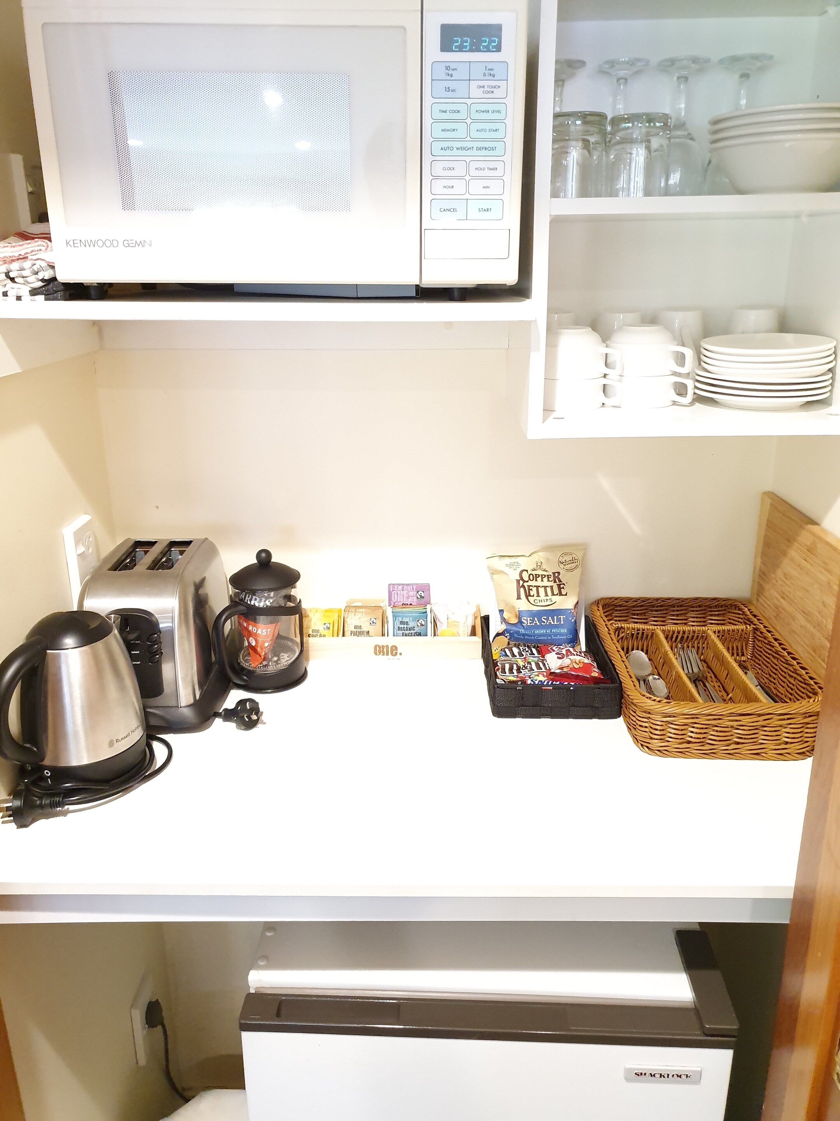 Una cocina equipada con microondas, tostadora, hervidor y cafetera. Hay estantes con vajilla y una cesta con utensilios.