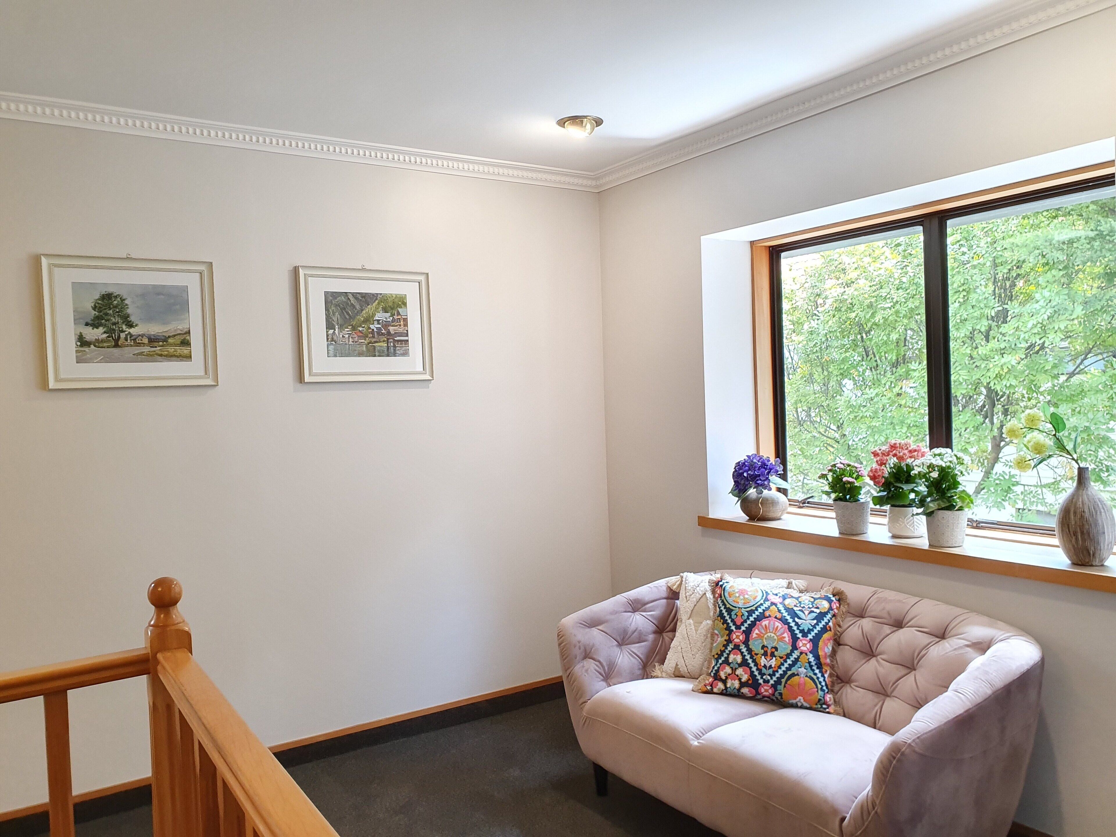 A room with a cushioned armchair, a window with a view of greenery, and two framed pictures on the wall.