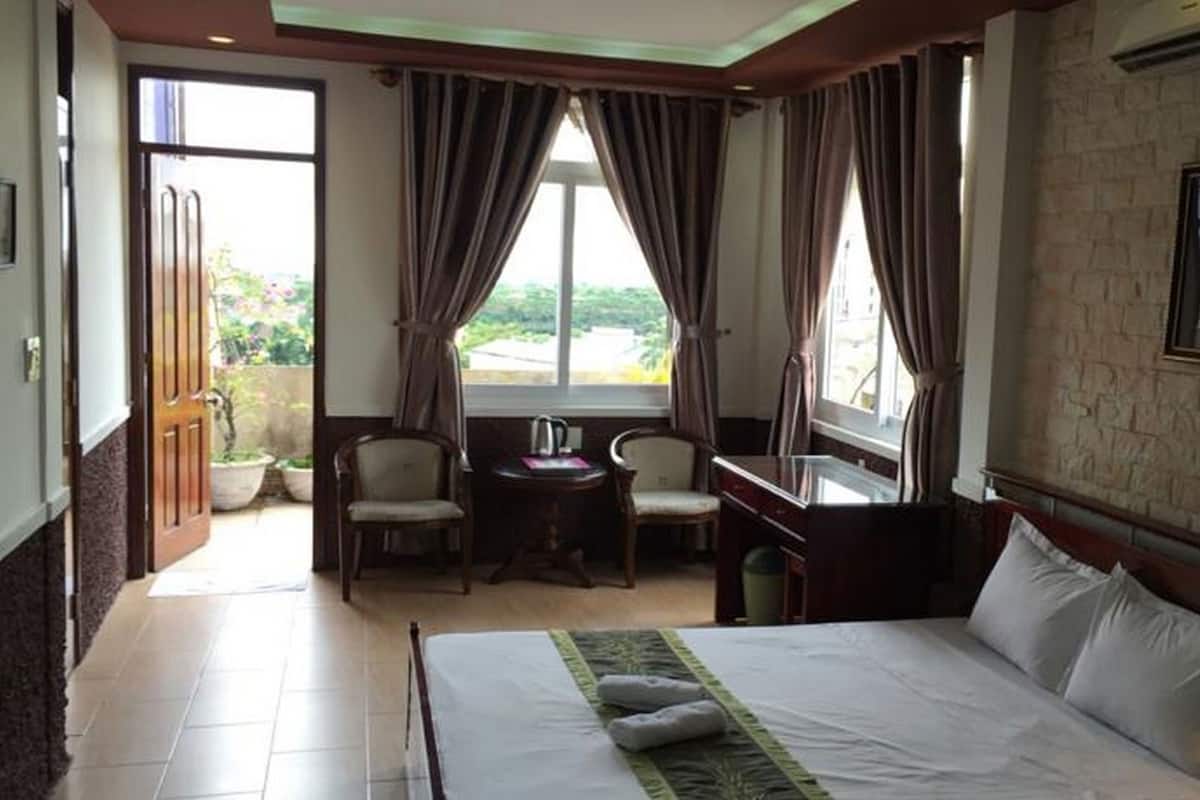 Habitación de hotel con cama, dos sillas, una mesita, ventana con cortinas y una lámpara.