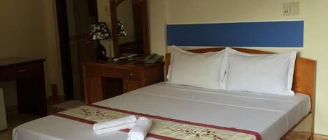 A hotel room with a wooden bed, white bedding, a desk with a lamp, and a mirror.
