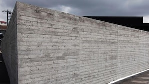 Paredes de concreto com padrão horizontal, calçada e um edifício ao fundo.