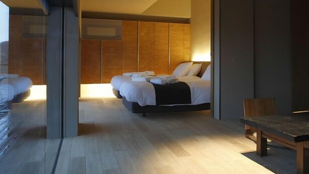 Quarto de hotel moderno com uma cama grande, cabeceira de madeira, uma mesinha e vista para a cidade.
