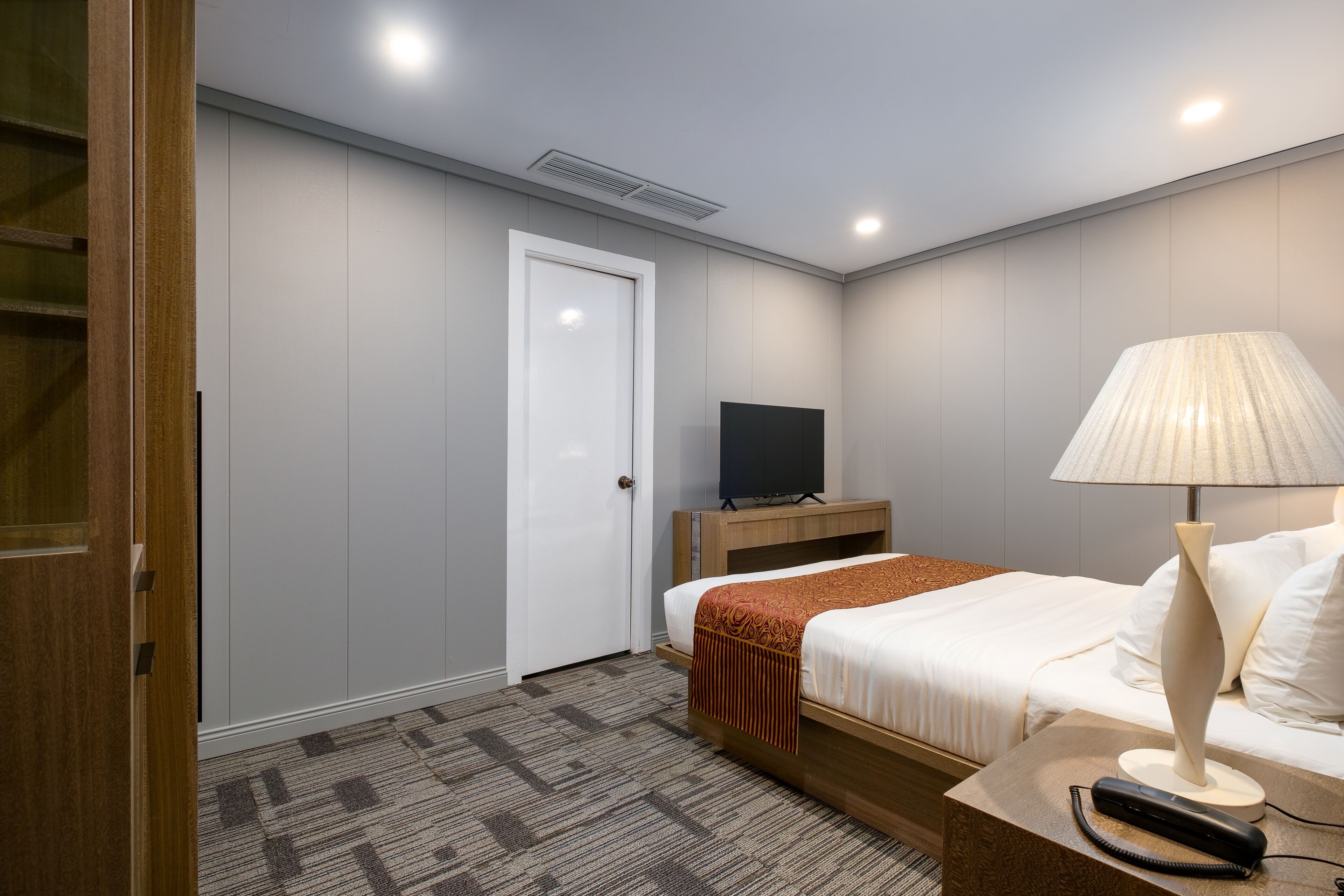 A hotel room with a bed, bedside table, lamp, TV, and a door.