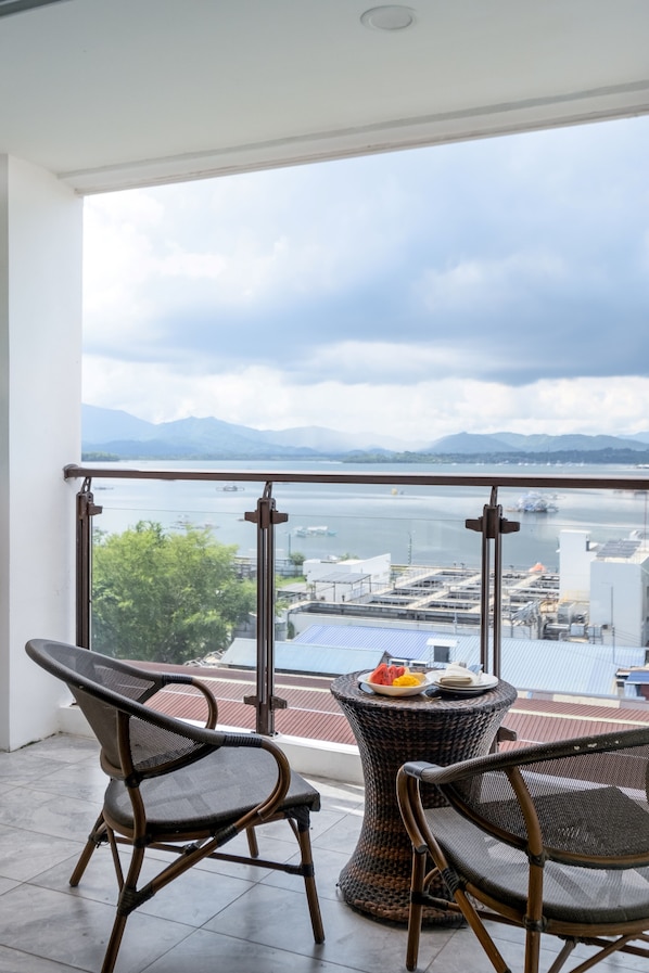 A balcony with wicker chairs and a table, overlooking a body of water and industrial buildings. - Sunlight Guest Hotel (Puerto Princesa)