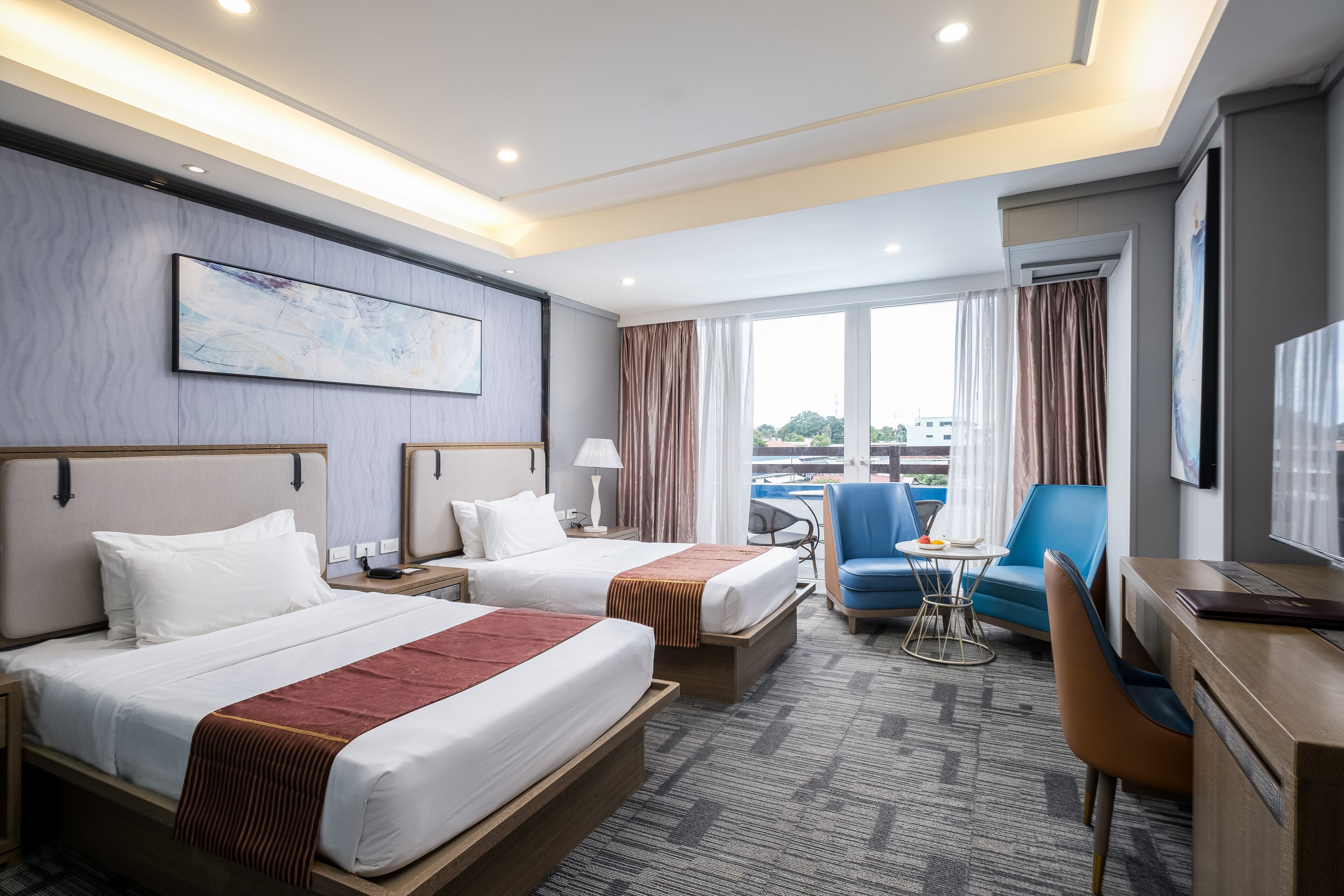Ein Hotelzimmer mit zwei Betten, einem Schreibtisch, einem Stuhl, einem kleinen Tisch und einem großen Fenster mit Vorhängen.