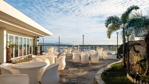 A rooftop terrace with white tables and chairs, a stone wall, and a view of the sea. - Sunlight Guest Hotel (Puerto Princesa)