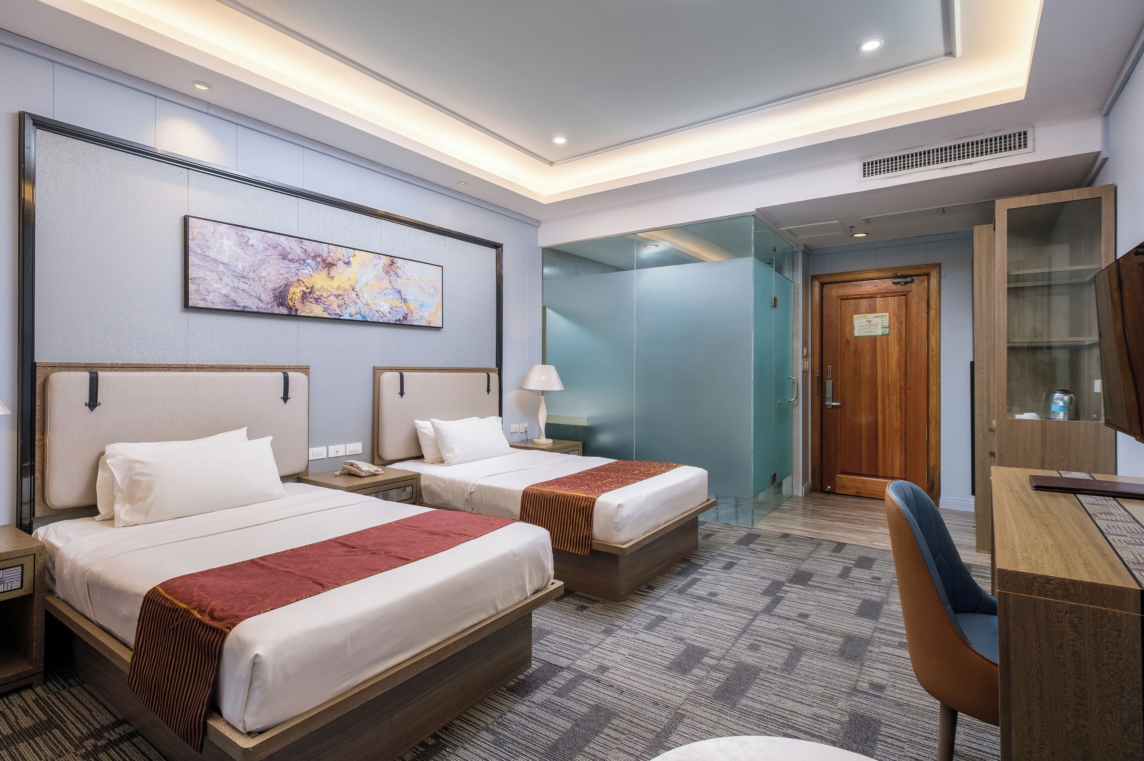 A hotel room with two beds, a desk, a chair, a large painting on the wall, and a glass partition.