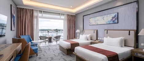A hotel room with two beds, a desk, and a chair. There is a view of the city through large windows.