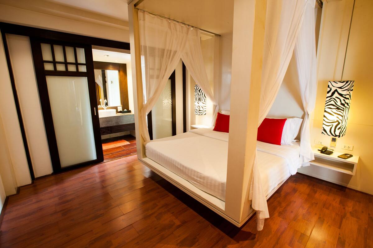 A bedroom with a four-poster bed, a nightstand with a lamp, and a wooden floor.