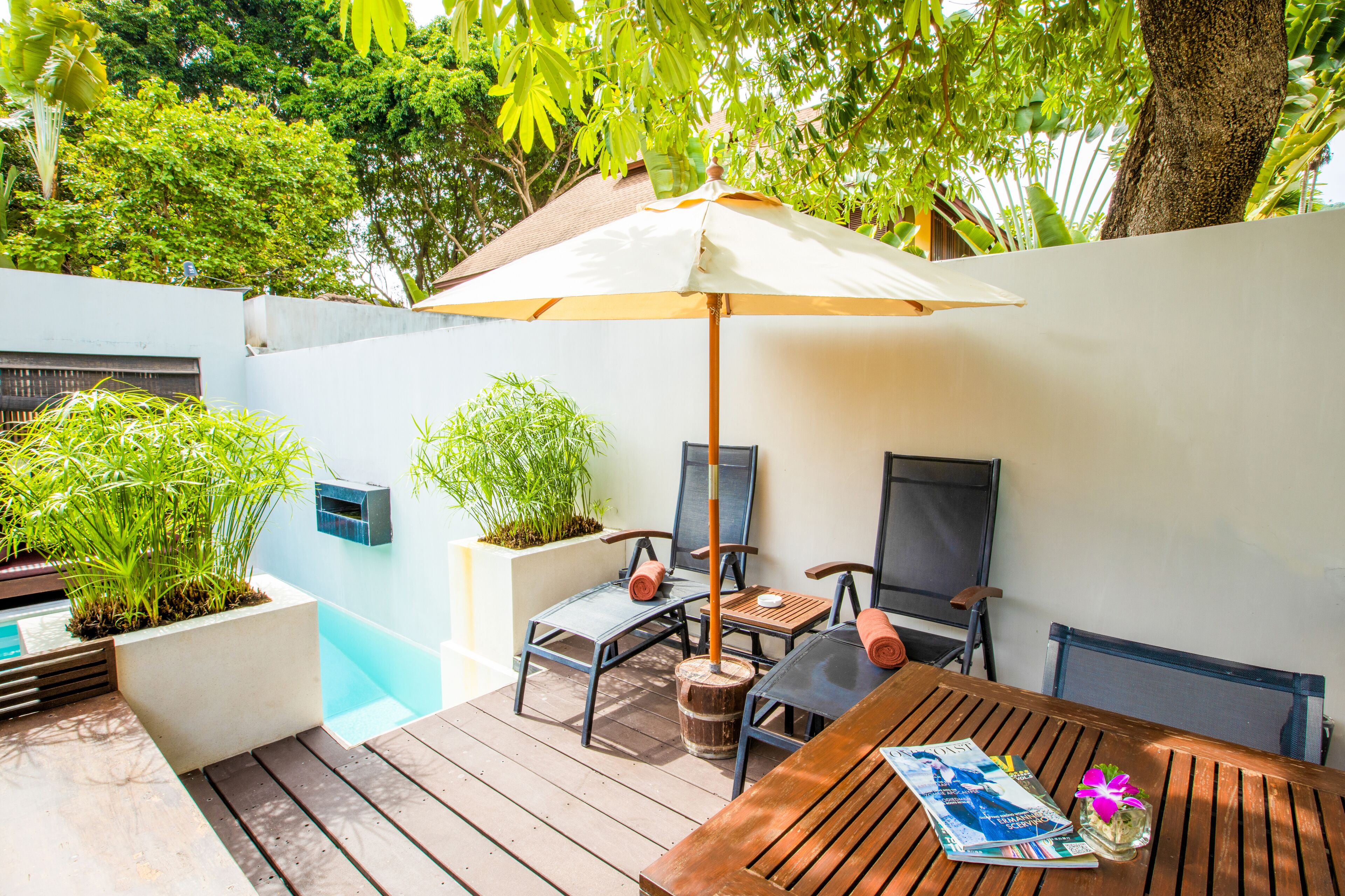 A wooden deck with a table and chairs under a large umbrella, surrounded by potted plants and a small pool.