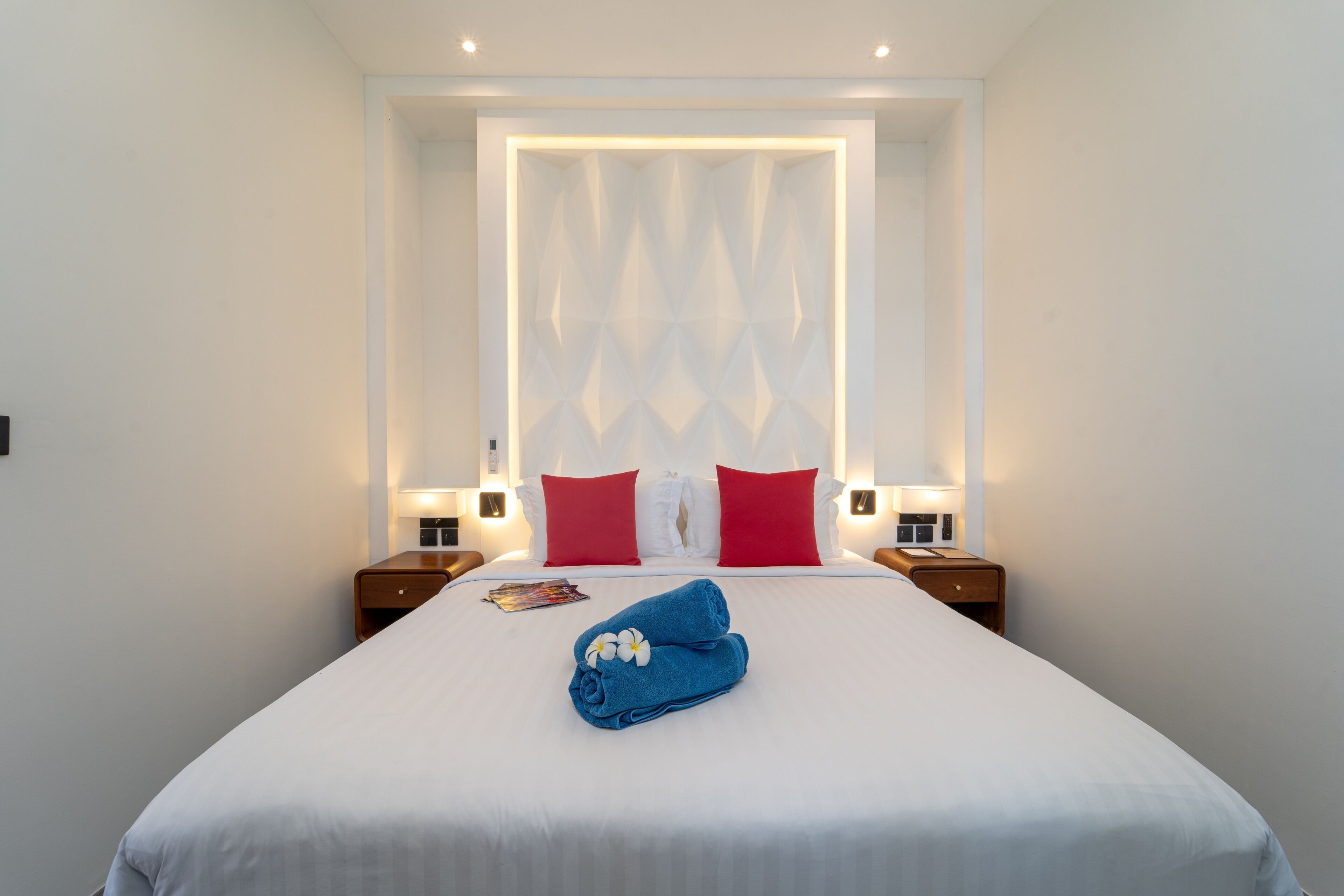 A neatly made bed with a white sheet, red and white pillows, and a blue towel with a flower. There are two bedside tables with lamps and a magazine on the bed.