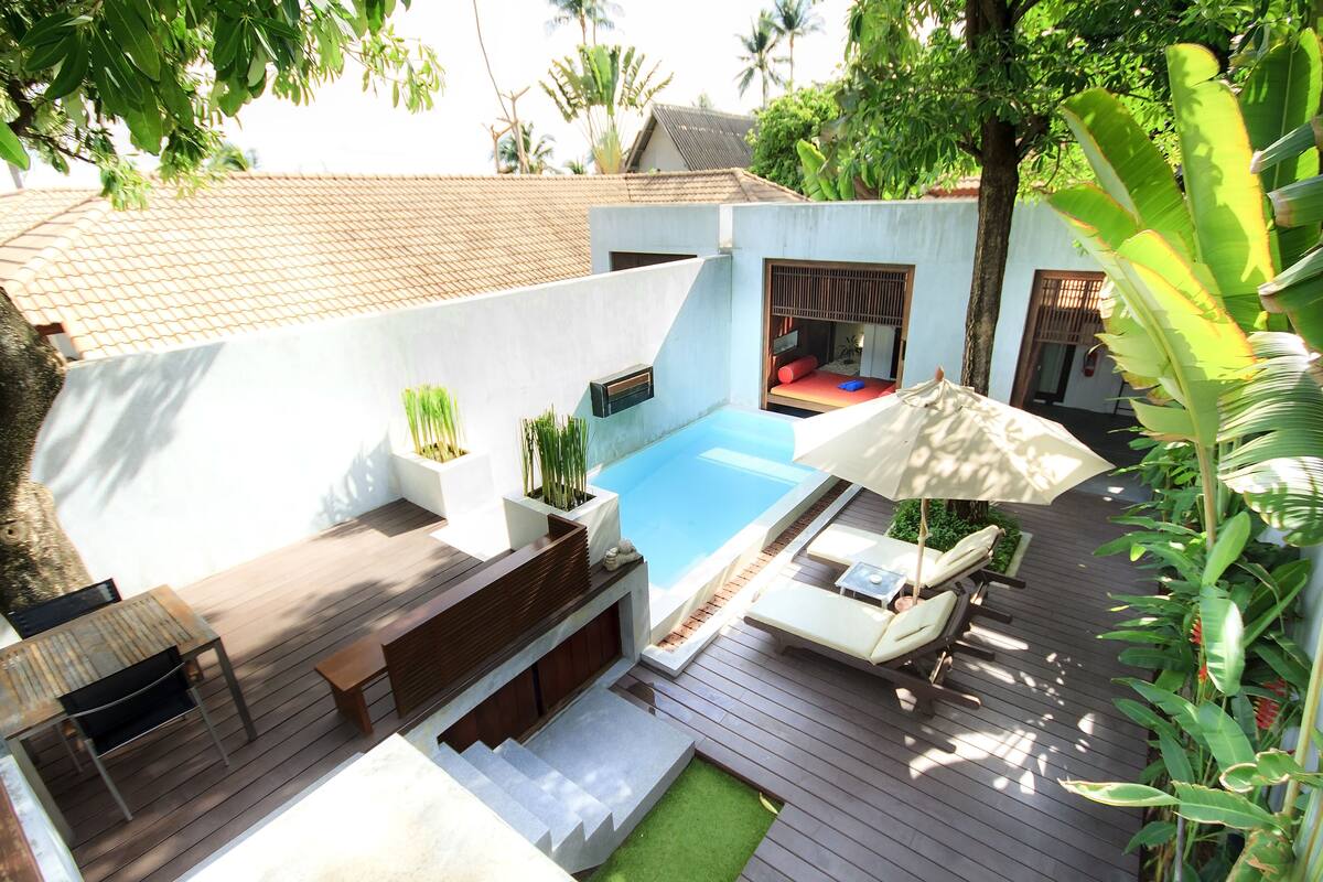 A poolside area with a wooden deck, a small seating area with a bench and a table, and a large umbrella providing shade.