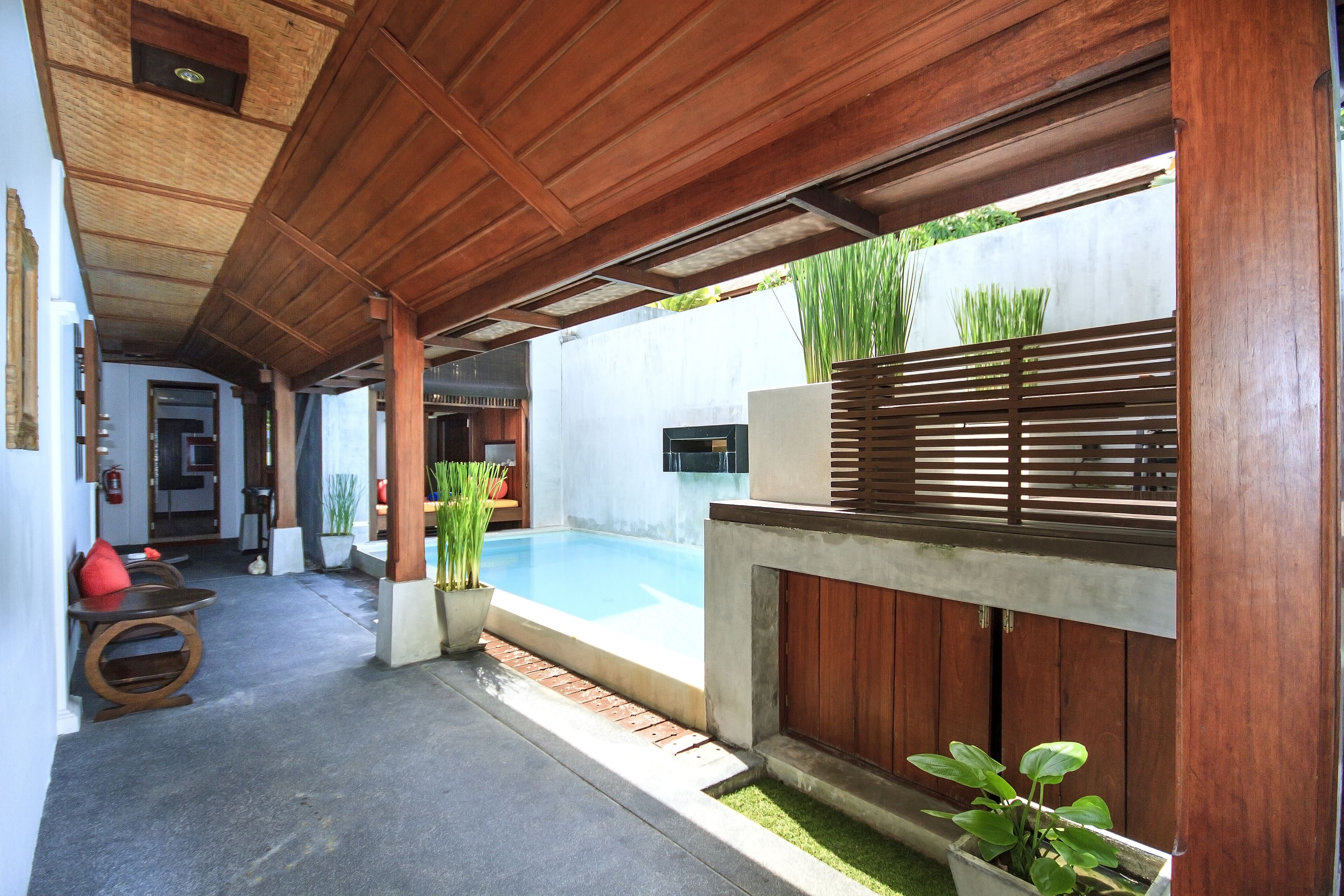 A covered outdoor area with a pool, wooden furniture, and potted plants.