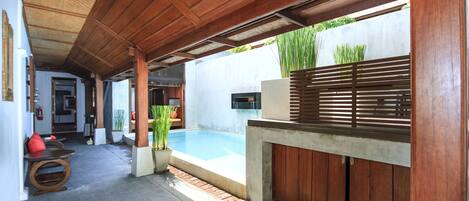 A covered outdoor area with a pool, wooden furniture, and potted plants.