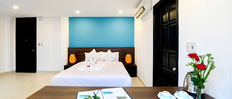A hotel room with a bed, a wooden nightstand, a table with a magazine, a vase with flowers, and a kettle.