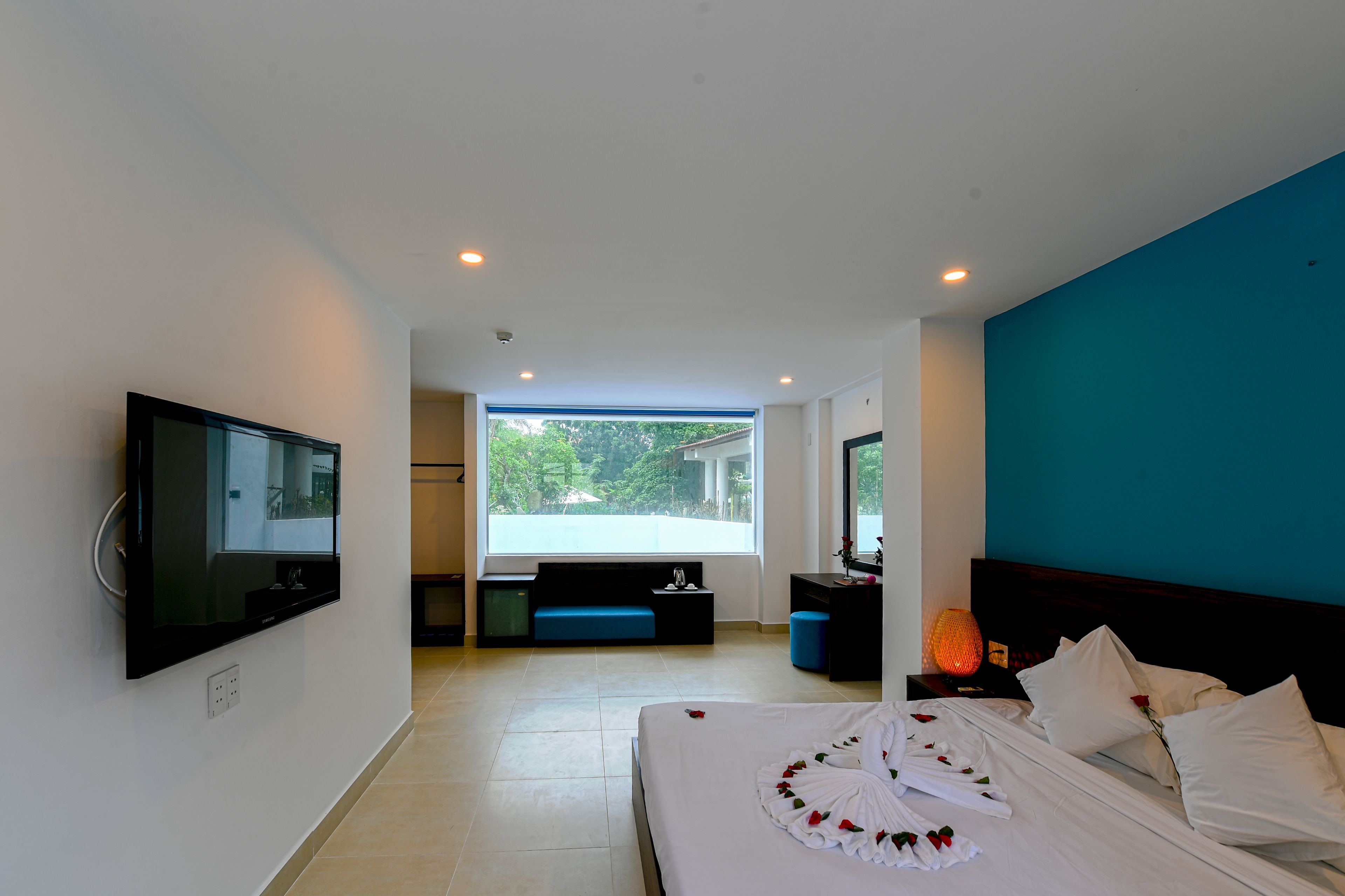 Una camera d'albergo moderna con un letto grande, una TV fissata al muro, una finestra con vista su vegetazione e una parete verde acqua.