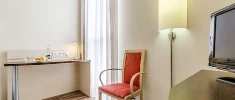 A hotel room with a wooden floor, a desk with a lamp, a chair, and a TV on a stand.